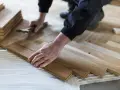 Ręce układają drewniane panele w jodełkę na kleju. Stos paneli czeka na montaż.