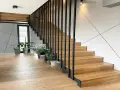 Nowoczesna klatka schodowa w domu z drewnianymi stopniami i czarną, metalową balustradą. Wokół rośliny w doniczkach.