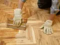 Ręce w rękawiczkach nakładają masę szpachlową na drewniany parkiet, pokazując, jak odnowić parkiet domowym sposobem.