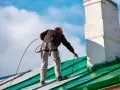 Pracownik na dachu wykonuje malowanie drewna pistoletem, odnawiając zielony dach pod błękitnym niebem.