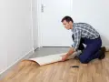 Mężczyzna w kombinezonie układa rolkę linoleum na podłodze. Pokazuje, jak położyć linoleum na płytki, przygotowując materiał do cięcia.
