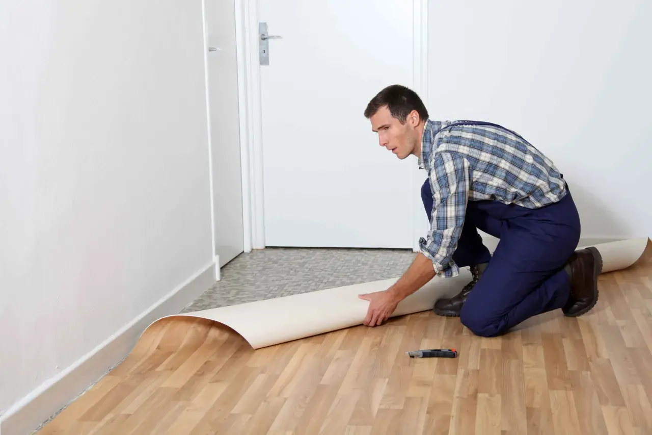 Mężczyzna w kombinezonie układa rolkę linoleum na podłodze. Pokazuje, jak położyć linoleum na płytki, przygotowując materiał do cięcia.