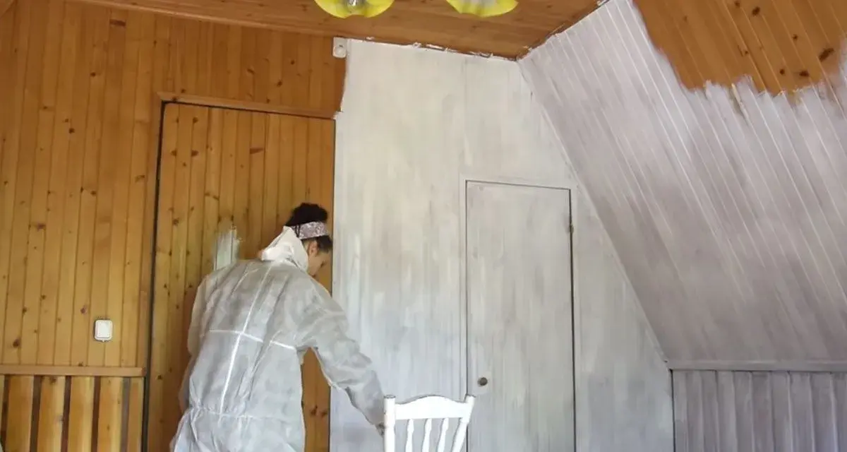 Osoba w kombinezonie maluje lakierowaną boazerię bez szlifowania na biało. Widać postęp prac na ścianie i suficie.