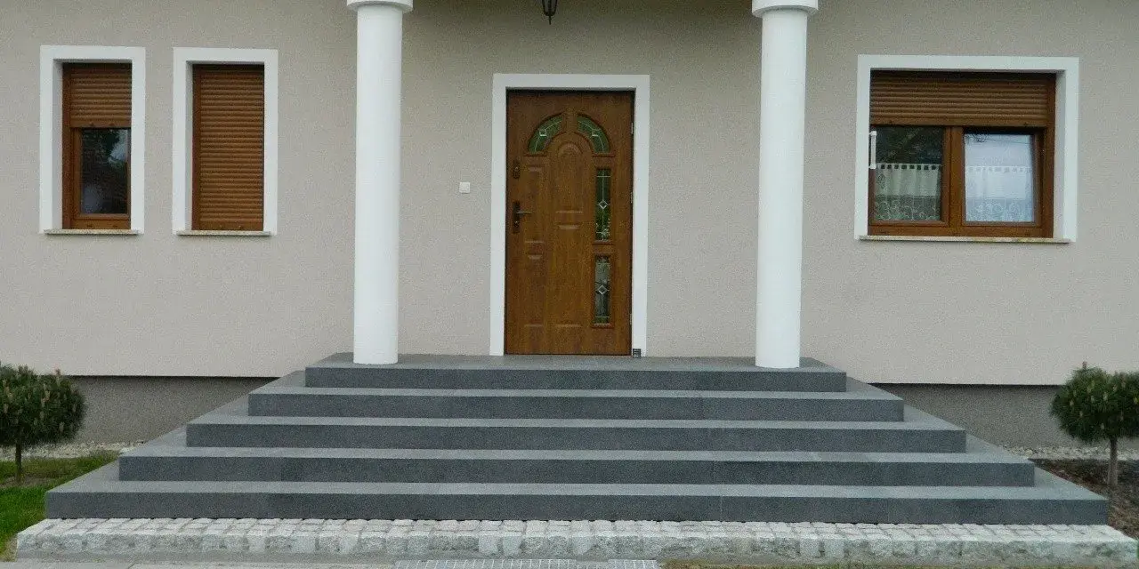 Eleganckie, szare schody zewnętrzne prowadzące do drewnianych drzwi. Świetny pomysł na schody zewnętrzne, podkreślający nowoczesny styl domu.