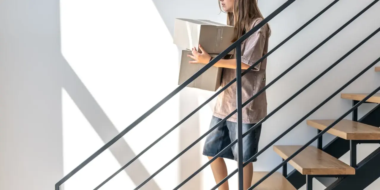 Nowa klatka schodowa w domu jednorodzinnym inspiracje: dziewczyna z kartonami schodzi po nowoczesnych schodach z drewnianymi stopniami i metalową balustradą.