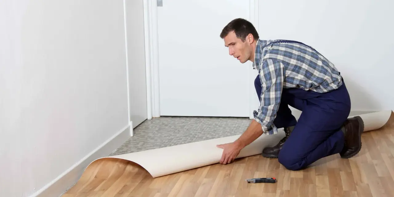 Mężczyzna w kombinezonie układa rolkę linoleum na podłodze. Pokazuje, jak położyć linoleum na płytki, przygotowując materiał do cięcia.