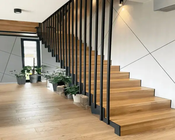 Nowoczesna klatka schodowa w domu z drewnianymi stopniami i czarną, metalową balustradą. Wokół rośliny w doniczkach.