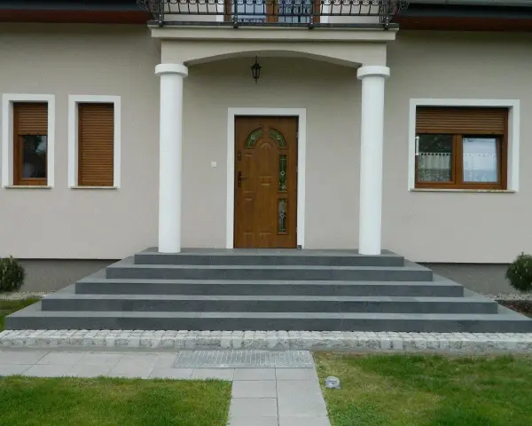 Eleganckie, szare schody zewnętrzne prowadzące do drewnianych drzwi. Świetny pomysł na schody zewnętrzne, podkreślający nowoczesny styl domu.
