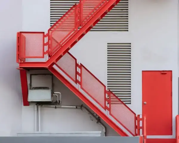 Nowoczesna, czerwona zabudowa schodów zewnętrznych z metalowymi stopniami i ażurowymi barierkami, prowadząca do czerwonych drzwi.