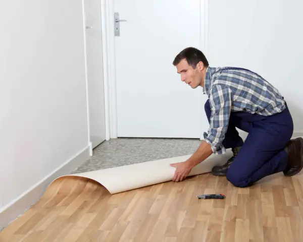 Mężczyzna w kombinezonie układa rolkę linoleum na podłodze. Pokazuje, jak położyć linoleum na płytki, przygotowując materiał do cięcia.