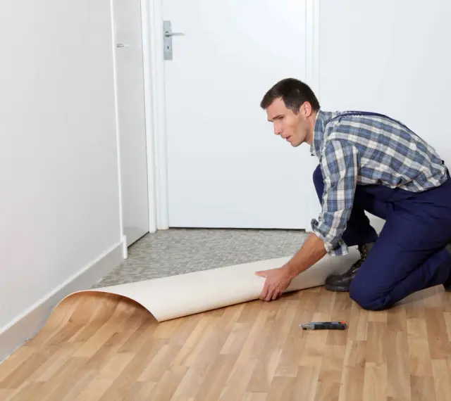 Mężczyzna w kombinezonie układa rolkę linoleum na podłodze. Pokazuje, jak położyć linoleum na płytki, przygotowując materiał do cięcia.