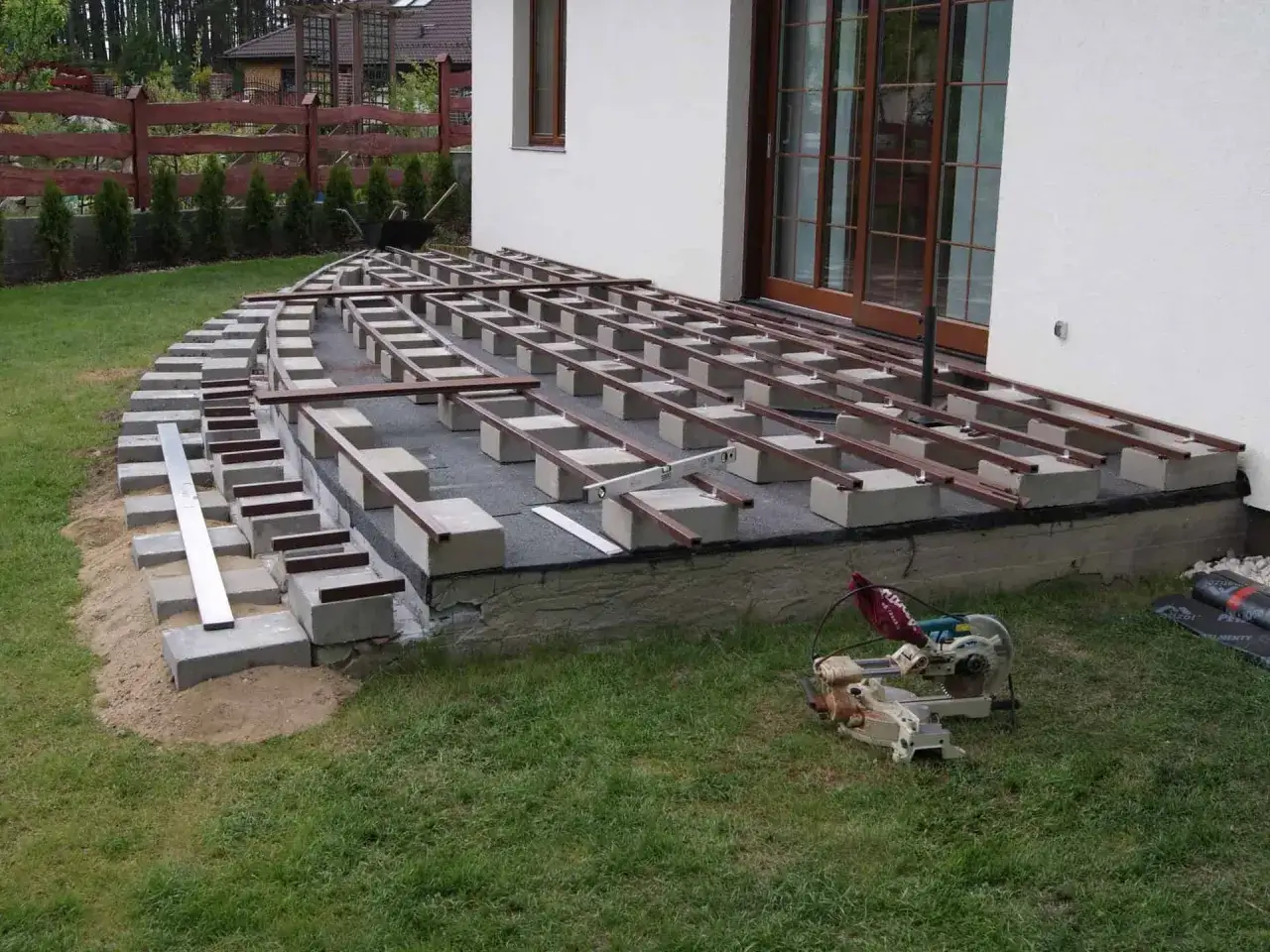 Rozpoczyna się montaż deski kompozytowej na betonowym fundamencie. Widać konstrukcję nośną z bloczków betonowych i legarów, gotową do ułożenia desek.