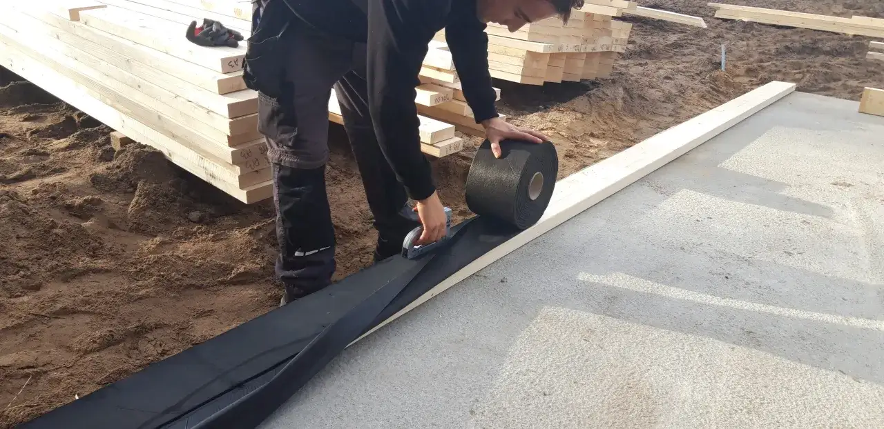 Pracownik układa czarną membranę, zapewniając izolację drewna od betonu. Na placu budowy leżą drewniane belki.
