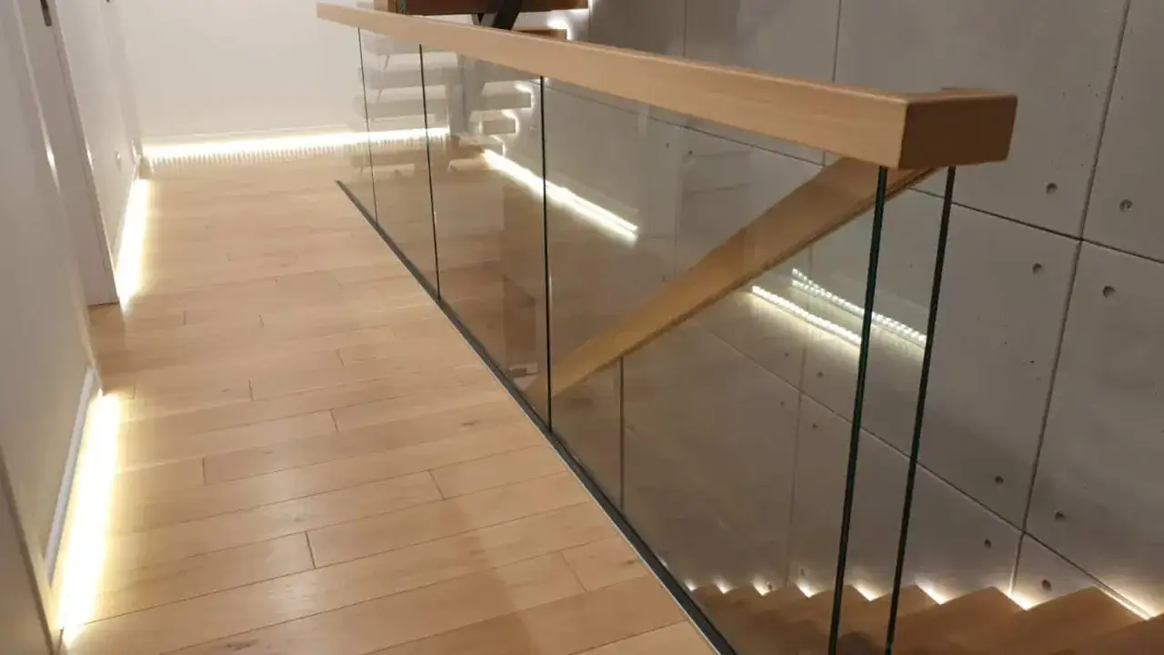 Nowoczesna klatka schodowa w domu z drewnianą poręczą i szklanymi balustradami. Podłoga z jasnego drewna i subtelne oświetlenie LED tworzą przytulną atmosferę.