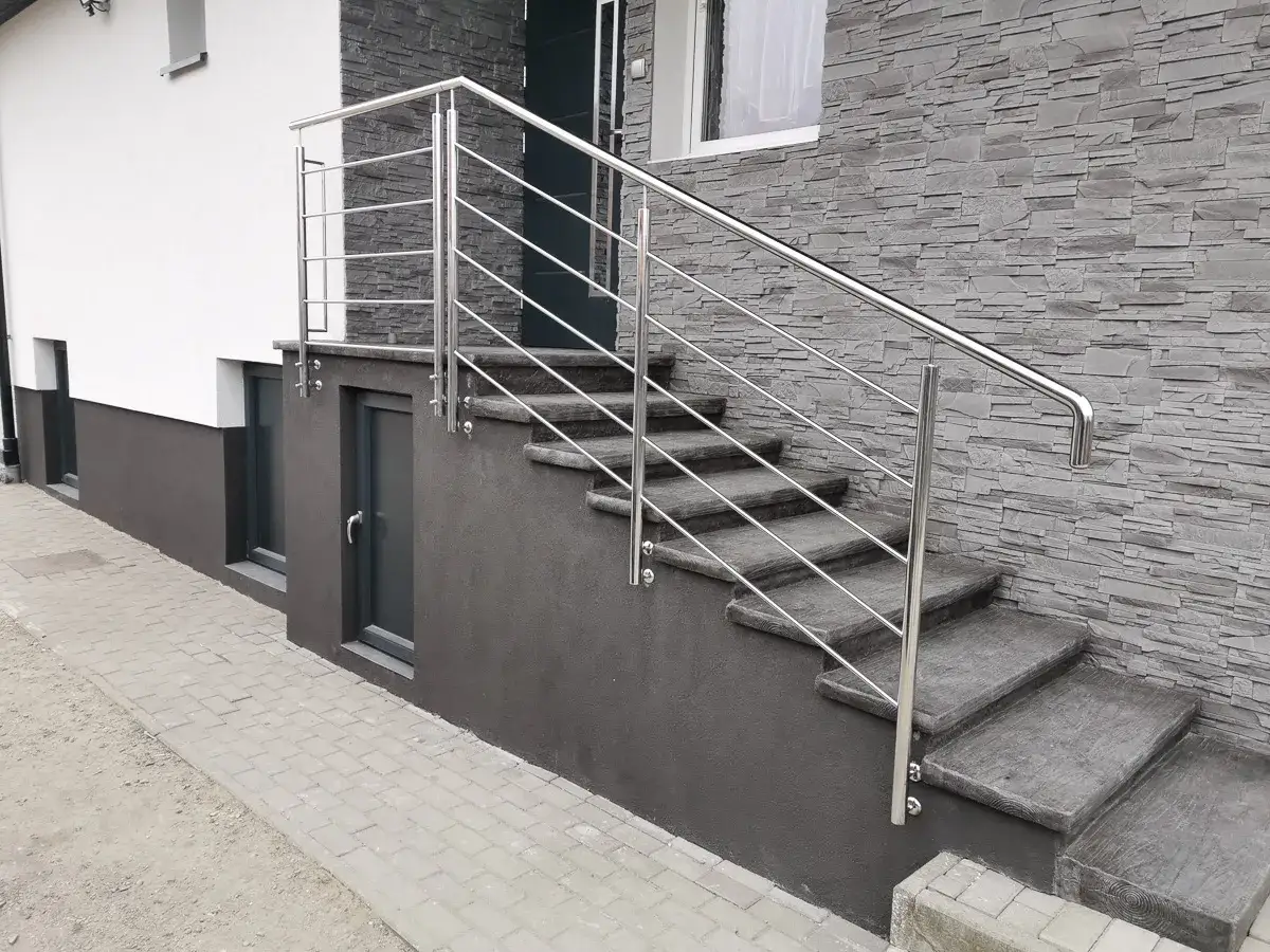 Nowoczesna, jaka balustrada na schody zewnętrzne ze stali nierdzewnej, idealnie komponuje się z kamienną elewacją i betonowymi stopniami.