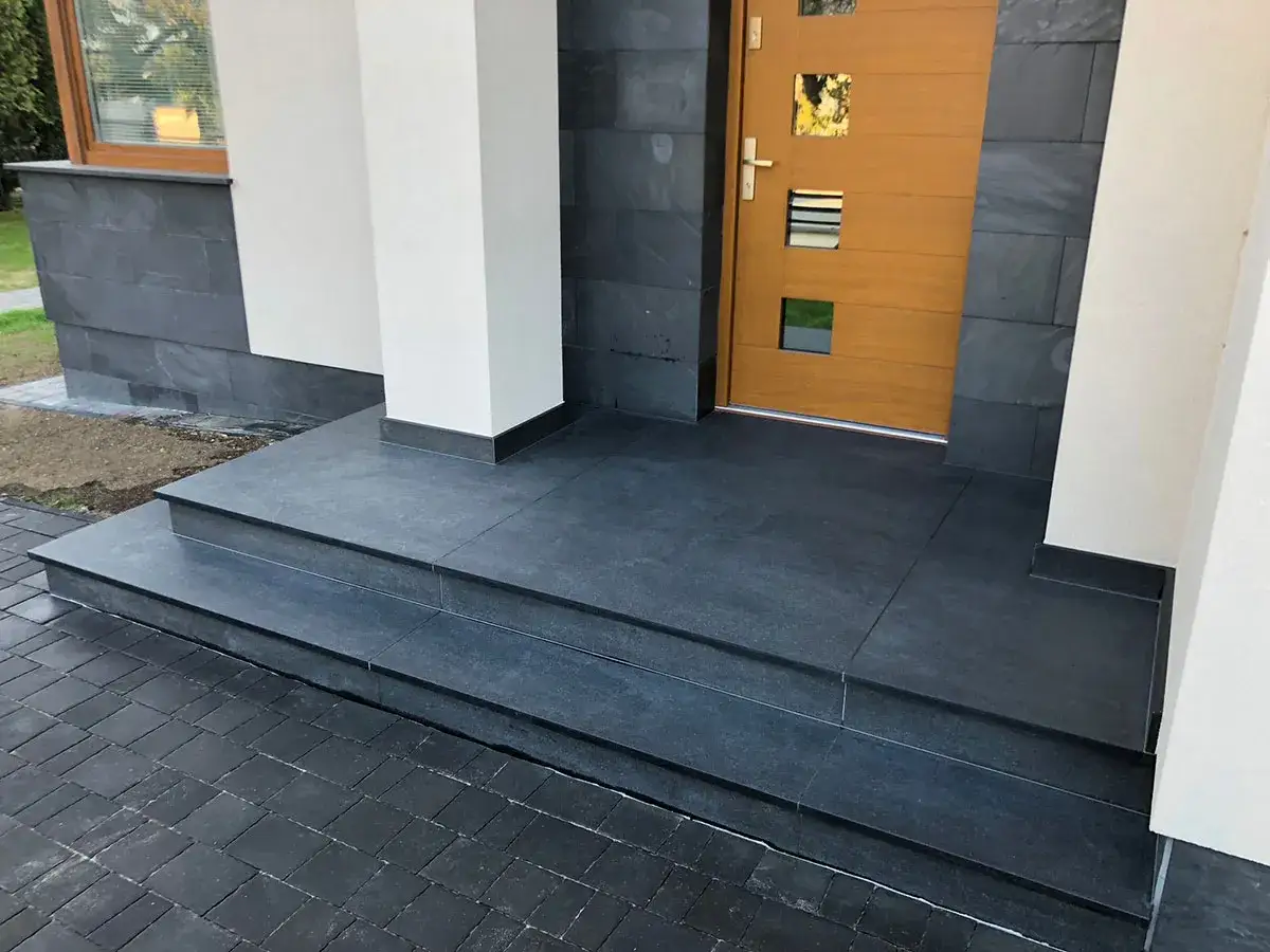 Nowoczesne schody zewnętrzne z ciemnoszarej płytek, idealny pomysł na schody zewnętrzne. Elegancko komponują się z drewnianymi drzwiami i kamienną elewacją.