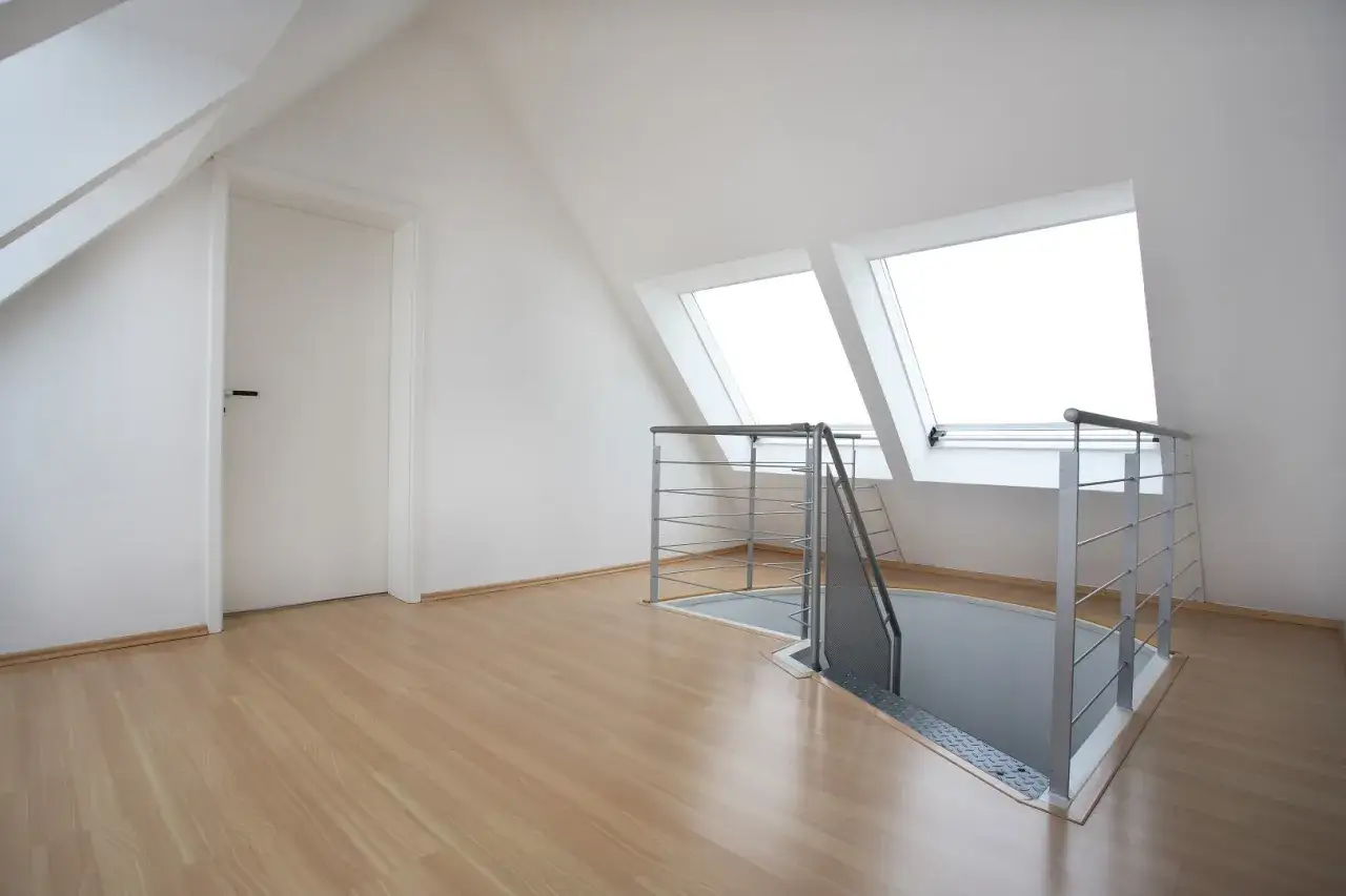Nowoczesne schody na poddasze z metalową balustradą prowadzą w dół. Jasne okna dachowe i drewniana podłoga tworzą przestronne wnętrze.