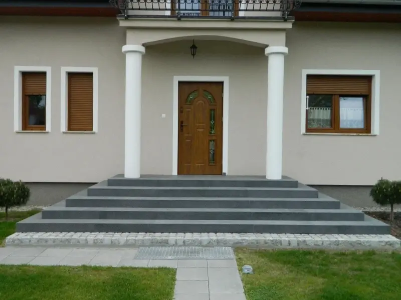 Eleganckie, szare schody zewnętrzne prowadzące do drewnianych drzwi. Świetny pomysł na schody zewnętrzne, podkreślający nowoczesny styl domu.